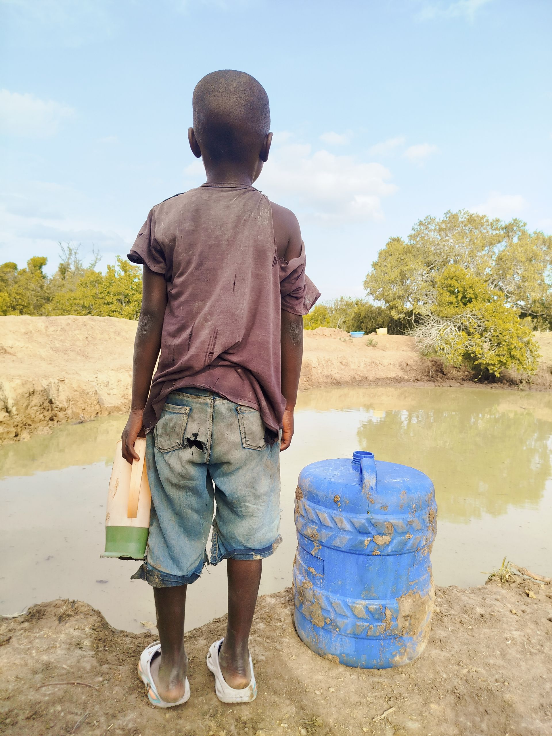 A young boy stands by a pond holding a container, symbolizing water scarcity.